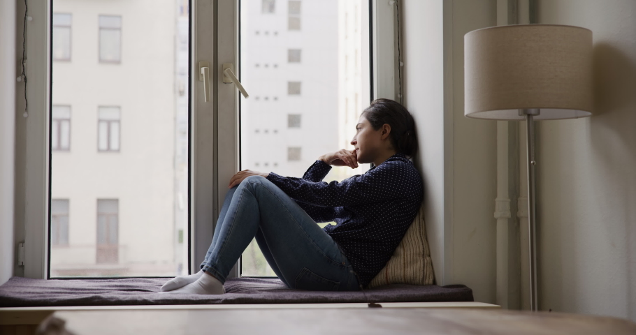 Melancholic Indian young woman sits on windowsill staring outside into window thinks over life concerns, feels upset. Frustrated female suffers from solitude looks sad. Relations break up, loneliness | Shutterstock HD Video #1096165049