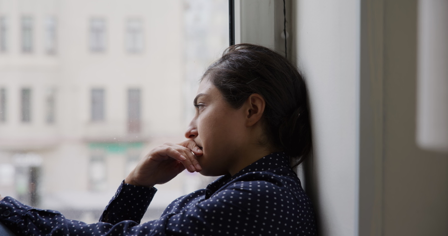 Close up upset young Indian woman sit on windowsill staring out window feels depressed deep in sad thoughts, looking concerned, due to personal troubles, break up, experiences life failure, melancholy Royalty-Free Stock Footage #1096165123