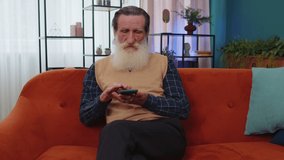 Senior grandfather sitting on sofa uses smartphone smile at home. Elderly man texting share messages content on social media applications online, watching relax movie, making shopping order delivery