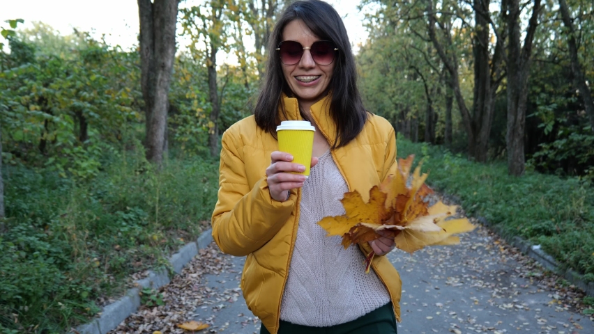 Young brunette woman in sunglasses drinks coffee with yellow autumn foliage. A woman in a yellow jacket is walking down the road. Slow motion portrait