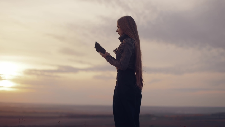 The woman closes the Bible, prays. A girl reads the Bible in the open air, studies the word of God at sunset on top of a mountain. Finding Truth in the Scriptures.