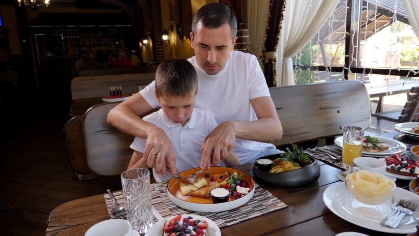 A young father and son have breakfast together in a cafe, dad cuts pancakes with a knife sitting at a wooden table. Royalty-Free Stock Footage #1096184645