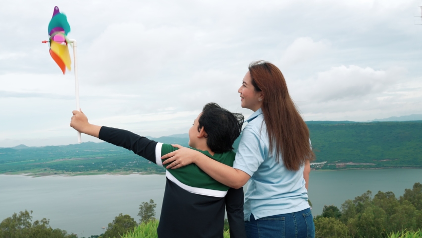 A progressive woman and her son are on vacation, enjoying the natural beauty of a lake at the bottom of a hill while the boy carries a toy windmill.