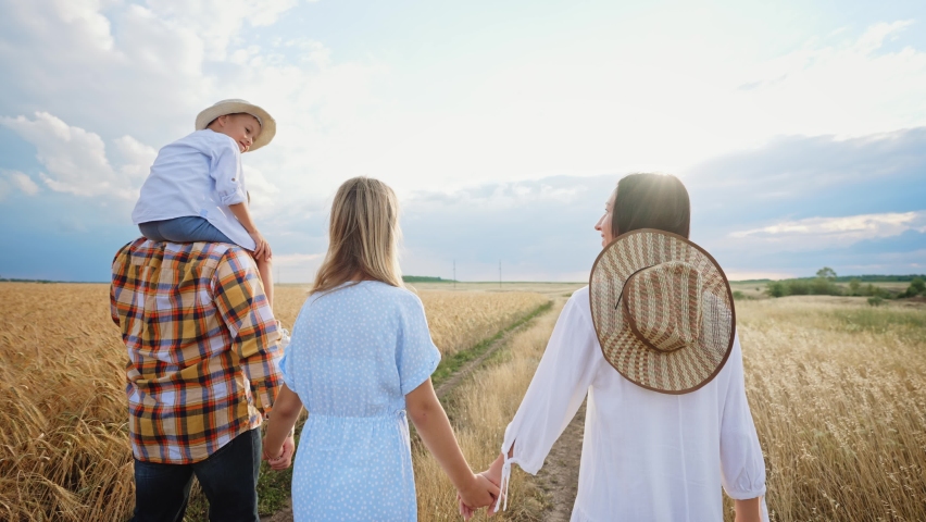 Rear view of family running on summer wheat field at sunset. Boy child sits on shoulders on his dad, holds his hands and they spread them apart. Mom and daughter in beautiful dresses walk in nature.
