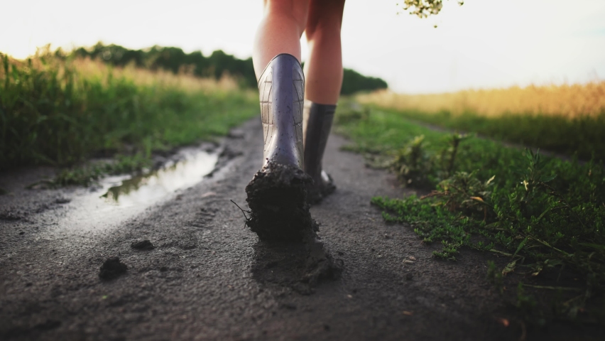 Rear view of legs of girl in dress and rubber boots who is walking along country road in field through wet puddles and ground after rain. Young woman holds bouquet of wildflowers in hand. Summer walk.