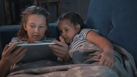 Two cute girls lying on couch under warm blanket in room are looking at screen of digital tablet.Modern digital entertainment technologies for children. Children learn through digital tablet, have fun