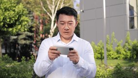Chinese handsome guy male urban professional businessman using mobile cell phone app in city to play video games online, enjoying free time, relaxing using smartphone, having fun excitement outdoor.