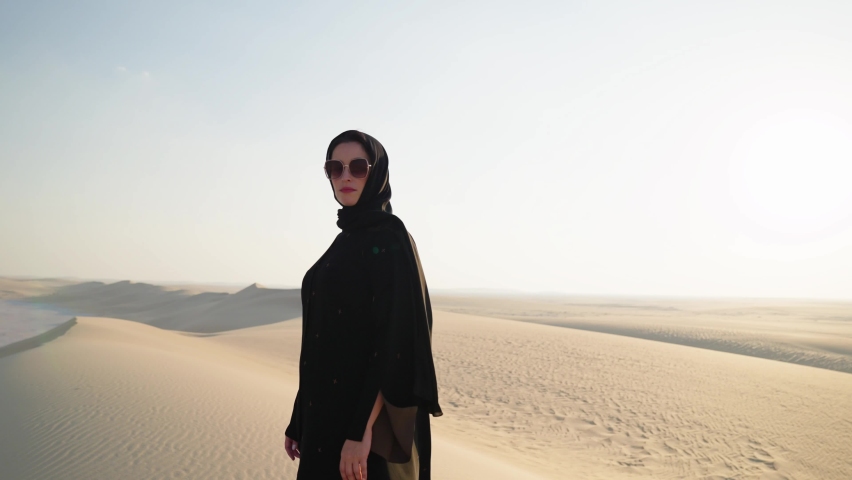Young beautiful girl in traditional Abaya dress seated on carpet along the dunes in the desert of Qatar

