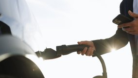 Unrecognizable businessman plugging electric car from charging station using app on smartphone. Man is plugging in power cord to an electric vehicle at sunset. Charging electric car. - Powered by Shutterstock - Get 15% off with code: PIKWIZARD15