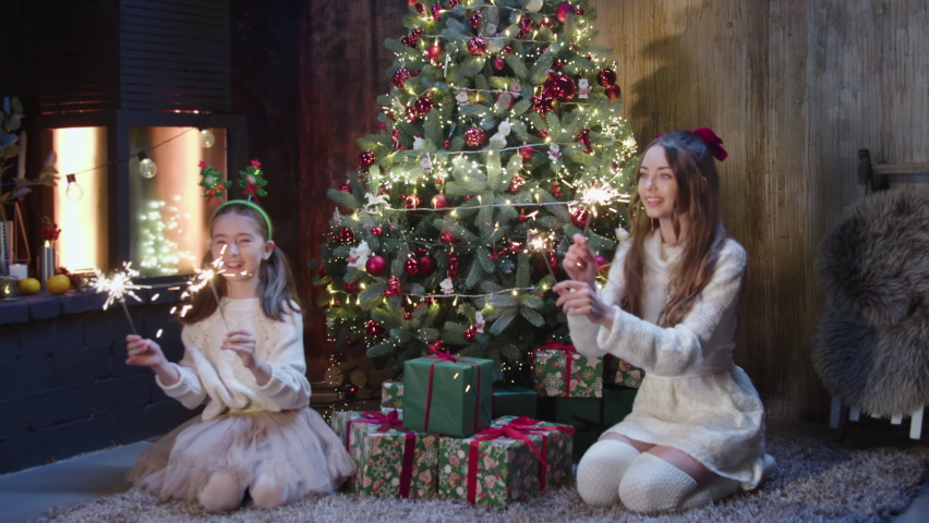 next to the holiday tree and next to a burning fireplace, children are sitting with burning sequins in their hands and enjoying the holiday. Two little girls are waiting for Santa Claus.