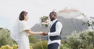 Video of happy diverse bride and groom holding hands, smiling and dancing at outdoor wedding. Marriage, love, happiness and inclusivity concept.