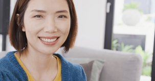 Video portrait of happy asian woman smiling to camera sitting at home. Domestic life, health, happiness and inclusivity concept.