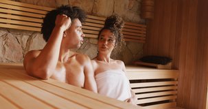 Video of relaxed diverse couple wearing towels sitting and talking in sauna room at health spa. Vacation, togetherness, relaxation, health and inclusivity concept.