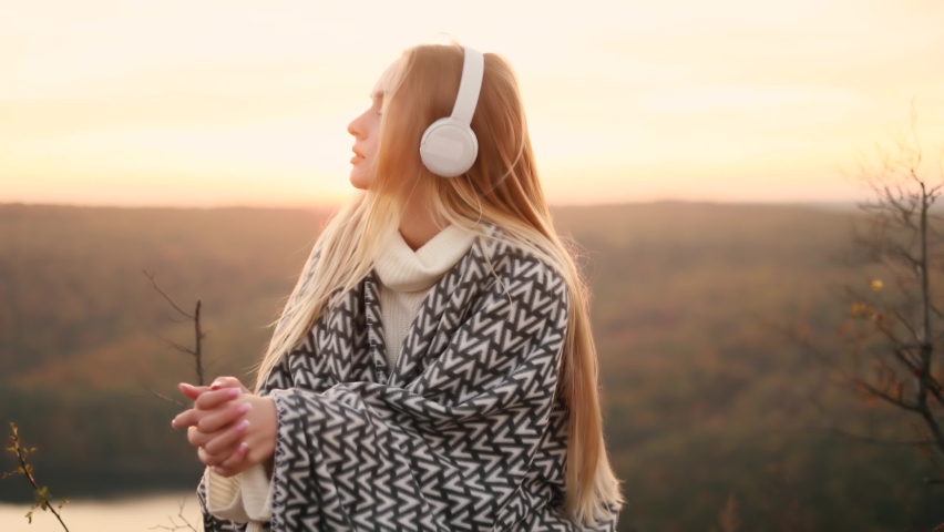 Relaxed young woman with long flying hair in headphones listening to music and dancing on the top of mountains with incredible sunset on the background. Inspiration time.