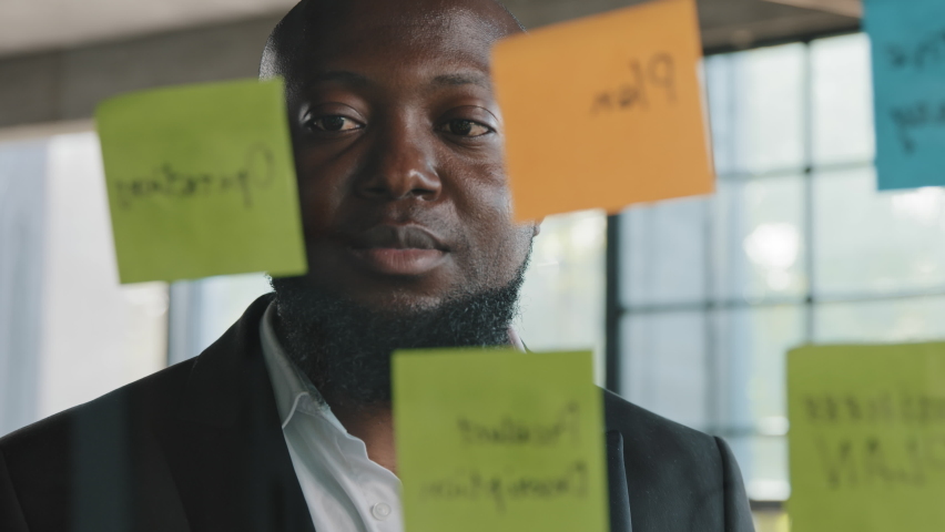 African American senior middle aged specialist businessman entrepreneur boss employer designer stand in modern office with glassy wall ethnic man worker read stickers notes organize management process