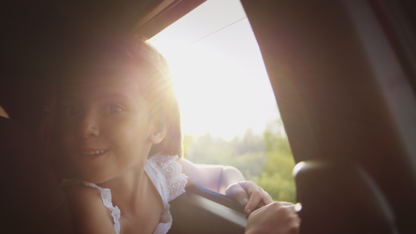 Cheerful little girl looks through open car window putting out hand and waving hello enjoying scenic landscape at sunset. Kid sits on backseat of automobile riding on road trip with family closeup Royalty-Free Stock Footage #1096226185