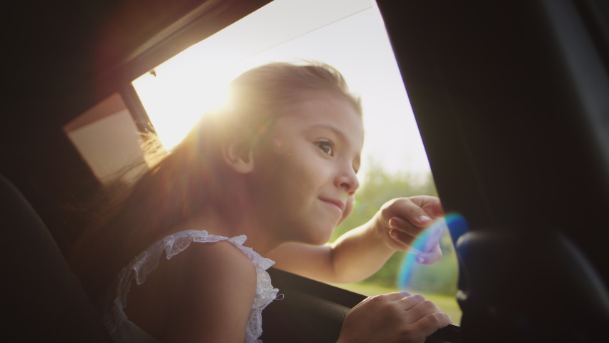 Cheerful little girl looks through open car window putting out hand and waving hello enjoying scenic landscape at sunset. Kid sits on backseat of automobile riding on road trip with family closeup | Shutterstock HD Video #1096226185
