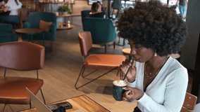 Happy African American woman sitting in cafe drinking coffee and working on laptop and phone, freelance businesswoman video call chatting typing message