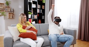 Happy joyful man playing dancing in virtual reality game enjoying music with beautiful woman on sofa. Cheerful relaxing male playing in VR goggles at home with surprised female. VR games concept.