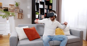 Happy relaxed man playing video game in virtual reality enjoying music. Beautiful joyful male dancing in VR helmet sitting on sofa at home, wearing sweater and jeans. VR games concept.
