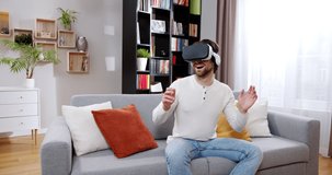 Adult beautiful man playing VR games sitting on sofa in living room. Stunning young male wearing VR headset waving, pointing hands, smiling and laughing. Virtual reality concept.