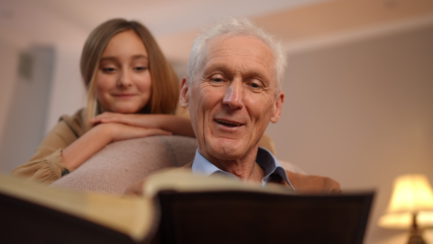 Portrait of Caucasian positive senior man talking recalling memories watching old photo album with blurred teenage girl listening stories at background. Happy grandfather and granddaughter at home