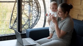 Young mother holds baby girl on lap sitting at table near window. Woman talks on video call via laptop showing screen to child with smile