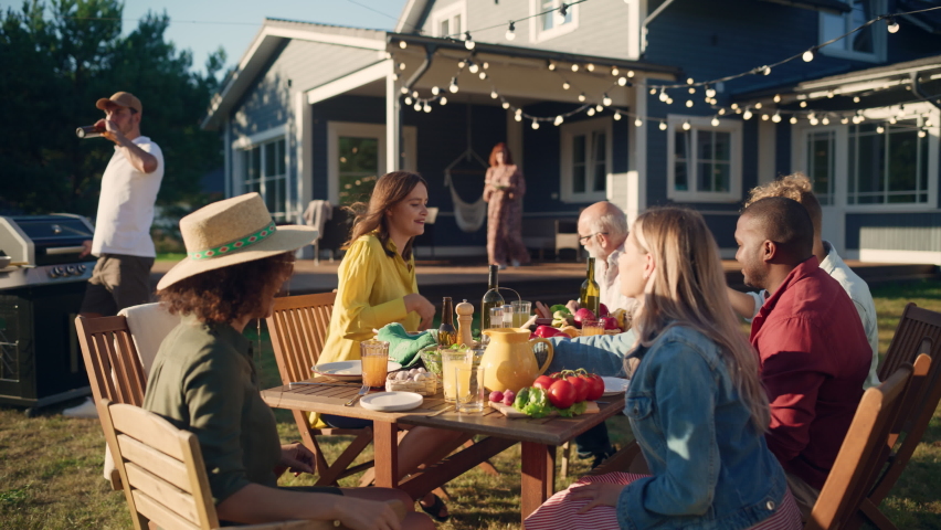 Parents, Children and Friends Gathered at a Barbecue Dinner Table Outside a Beautiful Home. Old and Young People Have Fun, Eat and Drink. Garden Party Celebration in a Backyard. Slow Motion Footage.