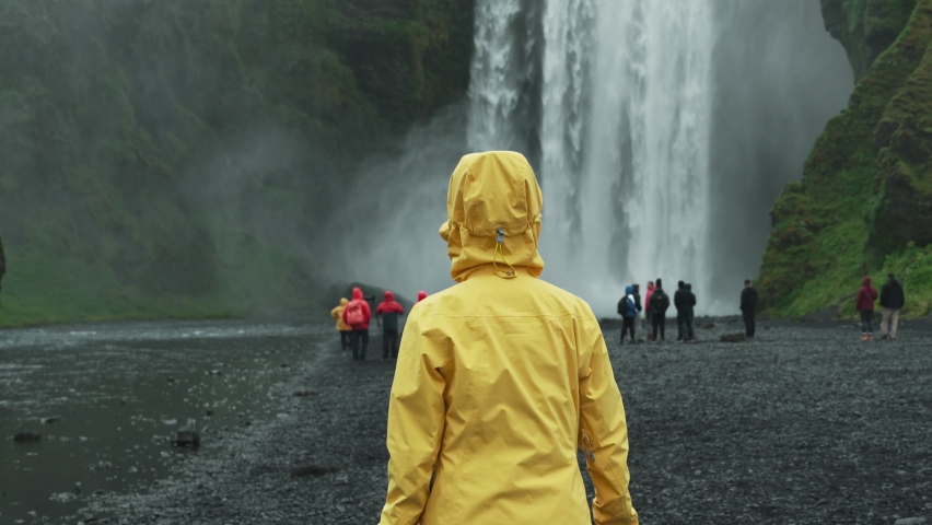 Female tourist in yellow jacket walking into powerful Skogafoss waterfall flowing in summer at Iceland