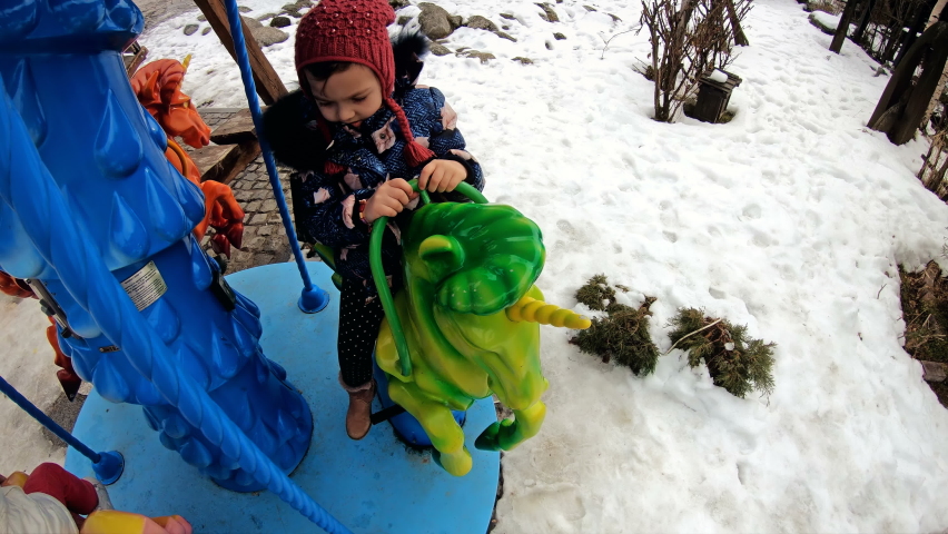 Happy little girl enjoying a ride on the merry-go-round outdoors on winter snow. Cute toddler kid having fun at funfair. Family with kids spending day in amusement park, slow motion