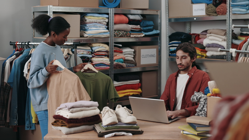 Multiracial female selecting clothes for recycle or donating and showing it to her male colleague at the charity shop. Reuse, second-hand, conscious consumer, sustainability concept Royalty-Free Stock Footage #1096260923