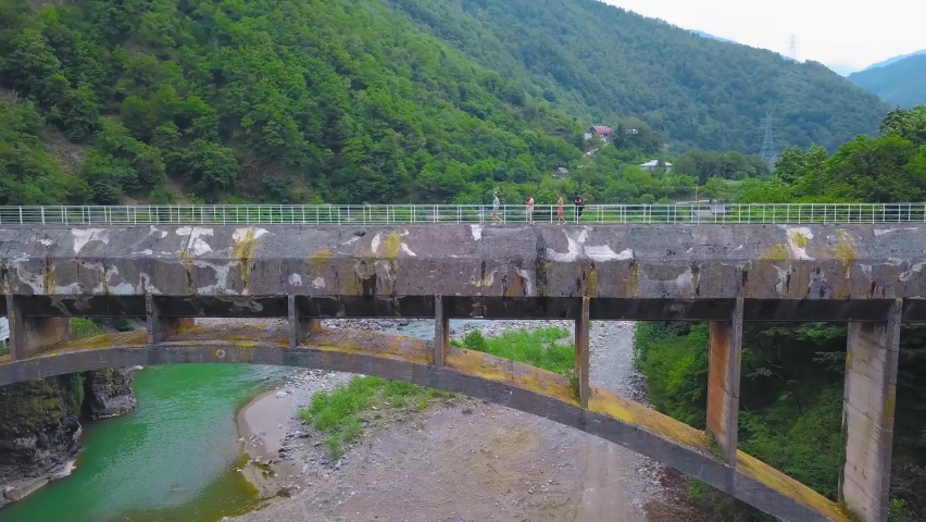 Company of four people walk across large concrete pedestrian bridge over gorge against backdrop of mountains and river in Georgia. Tourists explore area, landmark in form of ancient bridge drone.