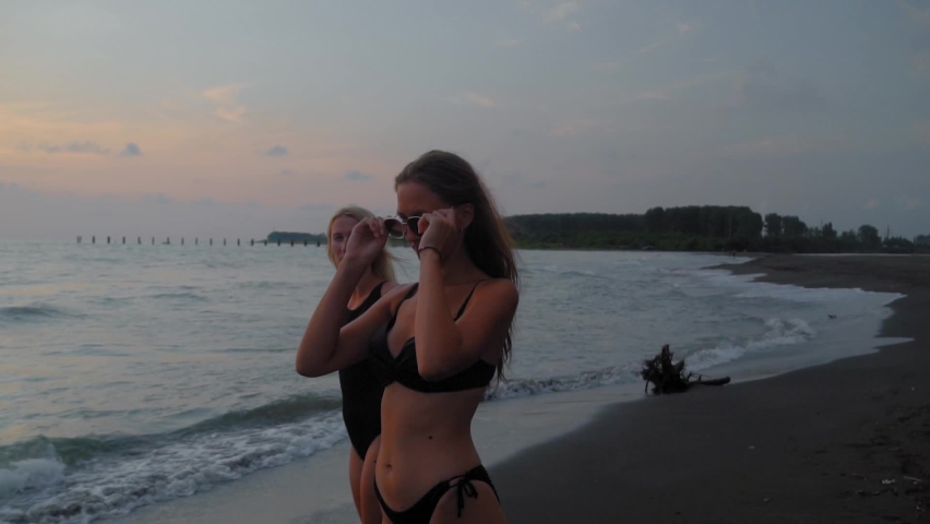 Two young beautiful women in black bikini walking along beach, along sea or ocean. stroll against background of waves and blue sky on horizon. Rest of two friends in sexy clothes. vacation on warm day