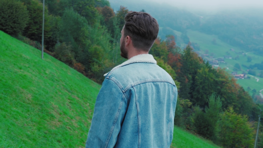 Back view of a guy walking in front of beautiful landscape with mountains and lake in Switzerland.  Bearded relaxed man male walking on a nature