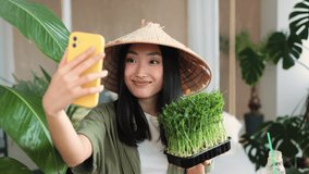 Beautiful asian woman blogger in traditional hat making vegetable fruit and microgreen sprouts salad and making video for social media enjoying healthy dinner in exotic light studio in tropical resort