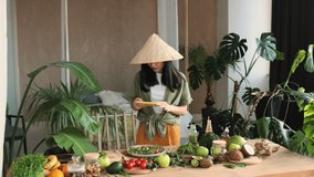 Young asian blogger woman using smartphone to posting photos in social media, standing in kitchen, female traditional conical hat making salad at exotic home. Lifestyle women relaxing at home concept.