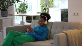 Young African American woman music lover lies on sofa with headphones on head and listens to songs from mobile phone playlist. Positive girl in casual clothes relaxes watching clips in smartphone 