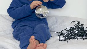 boy child preschooler opens gift cardboard box with christmas toys gray balls for fir tree.kid playing with rotating silver mirrored disco ball. blue color clothes lies on blanket undo christmas light