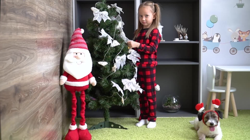 winter holidays and people concept - happy little girl decorating christmas tree at home. Nearby helps a pet dog in a funny Christmas hat of Santa Claus.