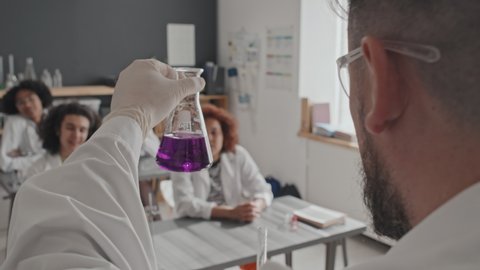 Over-shoulder of male chemistry teacher in medical gown and protective goggles standing in front of high school students in morning, mixing transparent and violet chemical liquids Video de stock