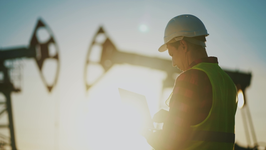 Oil business. a worker works next to an oil pump holding a laptop. industry business oil and gas concept. engineer studying the level of oil production on a laptop lifestyle silhouette at sunset