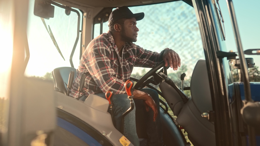Close up of happy African American young adult male farmer in cap sitting in tractor and smiling to camera. Positive man worker drives farm vehicle. Rural life. Agriculture farming business Royalty-Free Stock Footage #1096297053