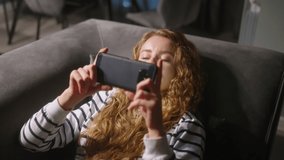 Woman scrolling smartphone, web surfing, lying on sofa at stylish living room at night. Female playing mobile games or shopping in store. Girl with phone enjoys chatting in social networks relaxing.