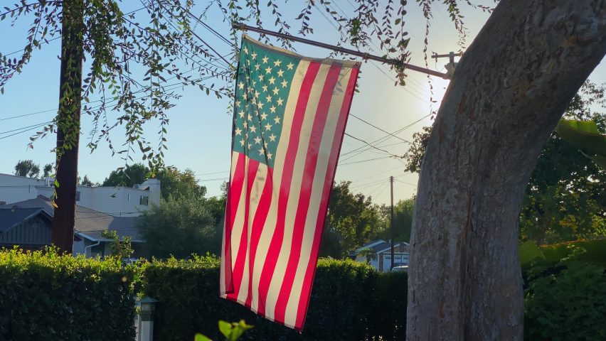 Shot of an American flag waving and swaying in the wind next to a tree in a residential area in a Neighborhood Area In Los Angeles USA, Blue Sky in Background, Establishing View