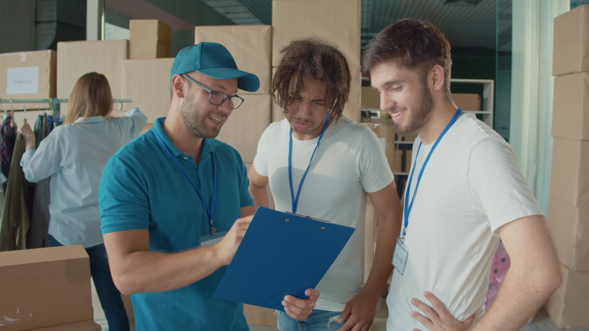 Male Volunteers at White T Shirts Talking With Mentor at Volunteer Warehouse. Charity, Donation, and Volunteering Concept. Work of the Volunteer Center Royalty-Free Stock Footage #1096311889