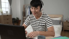 smiling asian student learning from home with headphones is nodding head to show understanding and taking notes while taking virtual lesson on laptop in bedroom