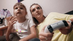 Funny young mom is playing video games on a console with her daughter holding a modern controller