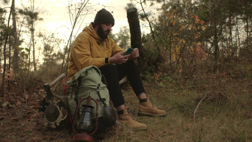 A bearded man rests on a fallen tree in the middle of the forest. A man uses a mobile phone to search GPS.