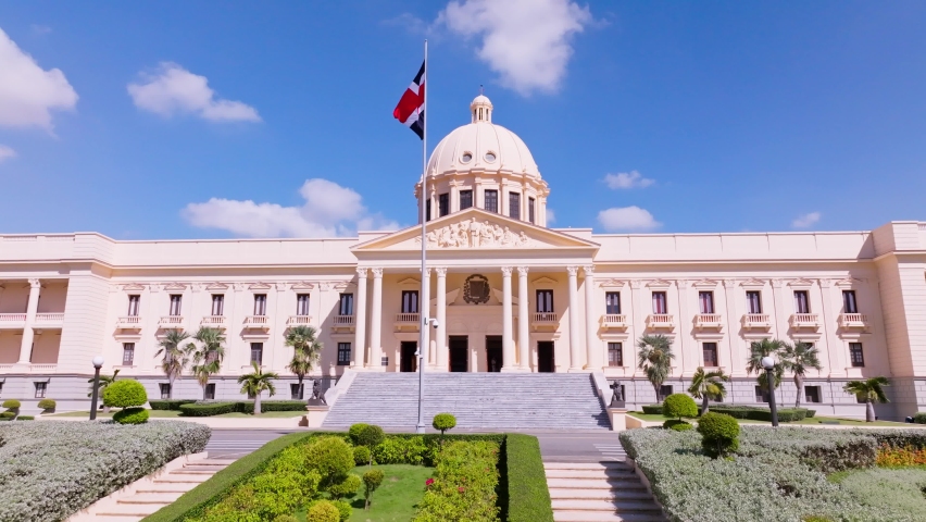 National Palace of Dominican Republic with flag waving in the wind against blue sky, Santo Domingo city. Aerial rising forward