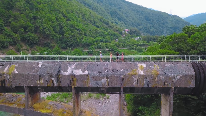 Company of four people walk across large concrete pedestrian bridge over gorge against backdrop of mountains and river in Georgia. Tourists explore area, landmark in form of ancient bridge drone.